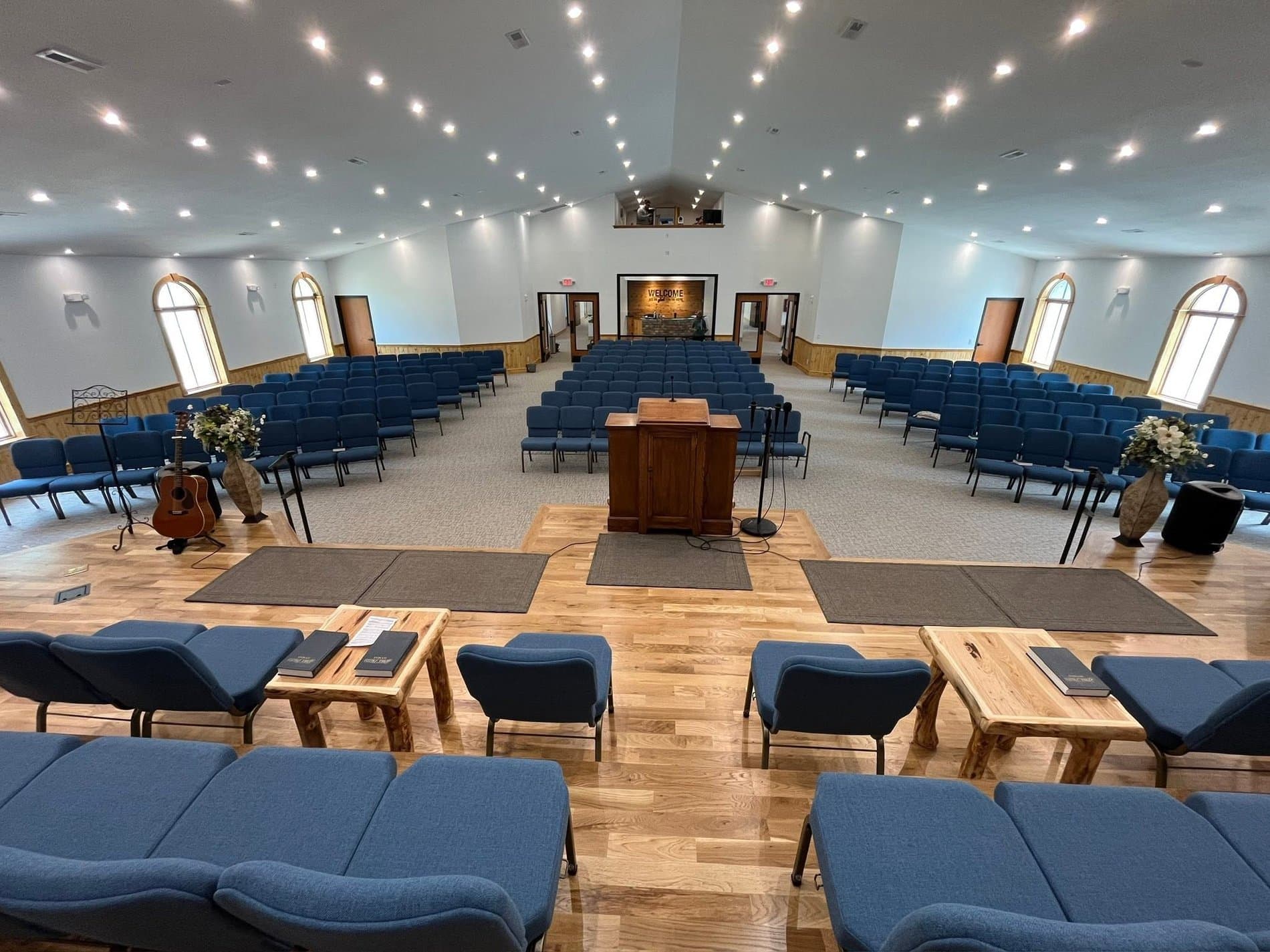 The auditorium at Calvary Baptist Church — rows of blue chairs facing a wooden pulpit, a stone cross on the platform wall, arched windows down both sides, and a wood floor